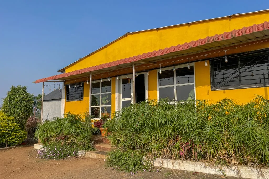 A vibrant yellow building with glass rooms and large windows, surrounded by lush greenery; located near Pawna Lake under a clear blue sky.