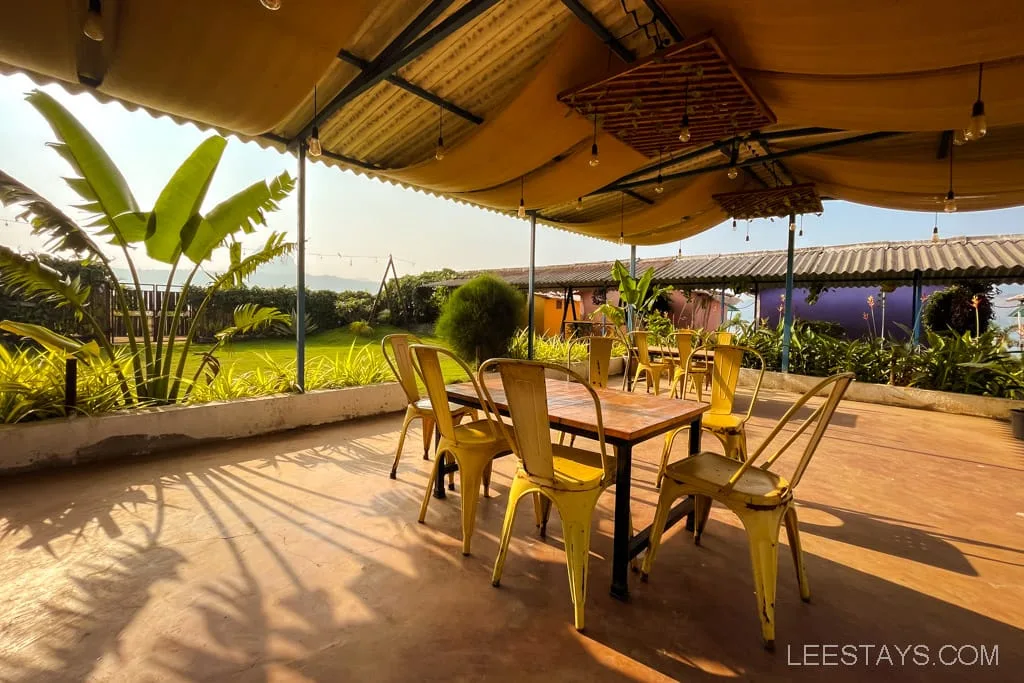 Outdoor seating area with yellow chairs and tables under a canopy, surrounded by greenery near glass rooms with a view of Pawna Lake in the background.