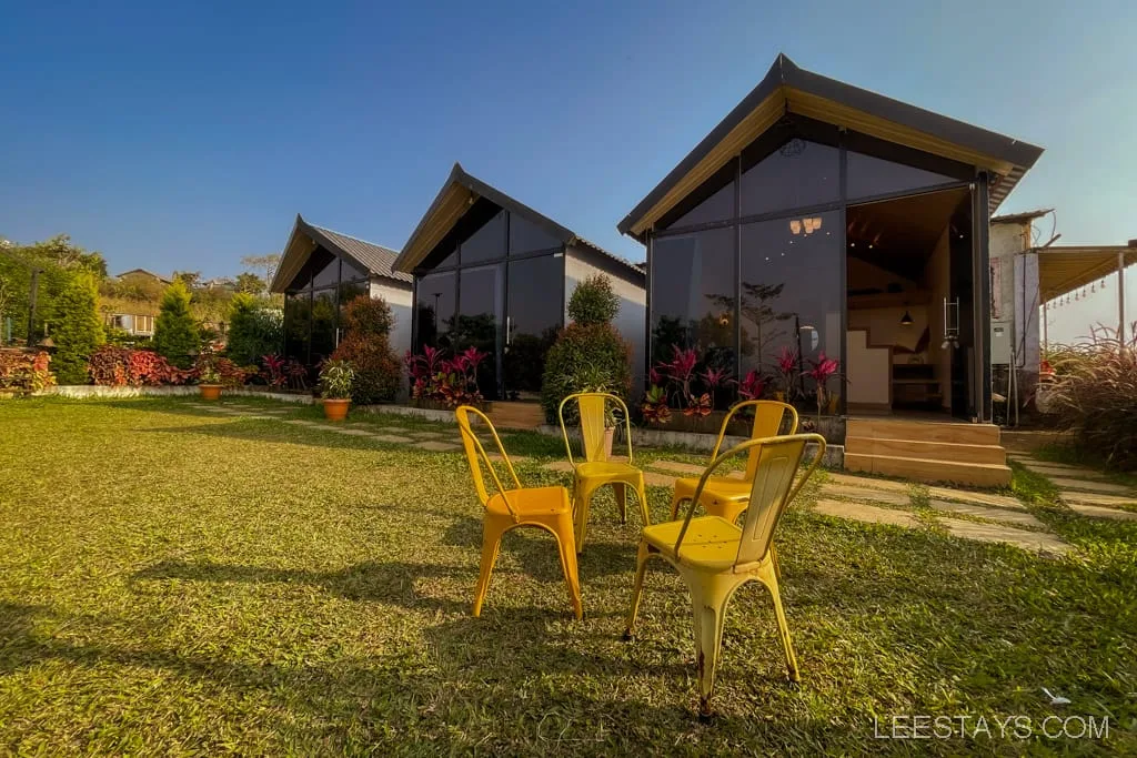 Scenic view of two glass rooms near Pawna Lake, surrounded by plants and a lawn with yellow chairs, under a clear blue sky.