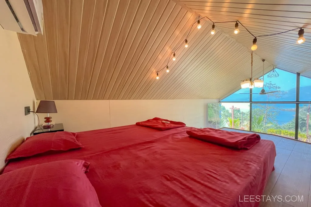 A cozy glass room at Pawna Lake with large windows, featuring a king-sized bed with red linens, string lights, and a view of the lake and greenery.