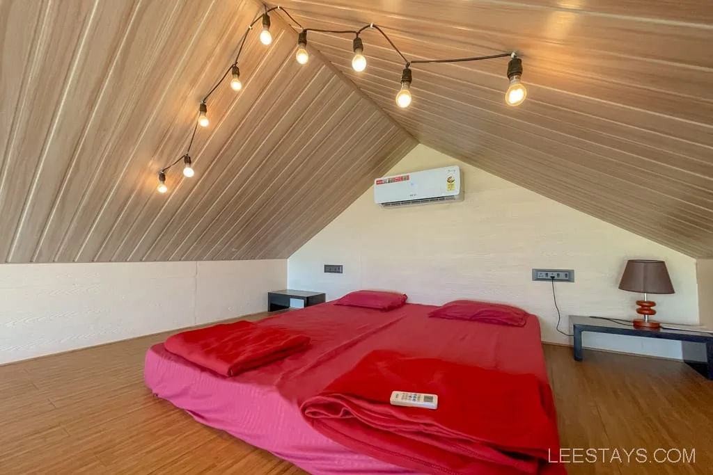 Cozy attic bedroom with ambient string lights, air conditioning, and a neatly made bed with red linens, reminiscent of accommodations near Pawna Lake's glass rooms.