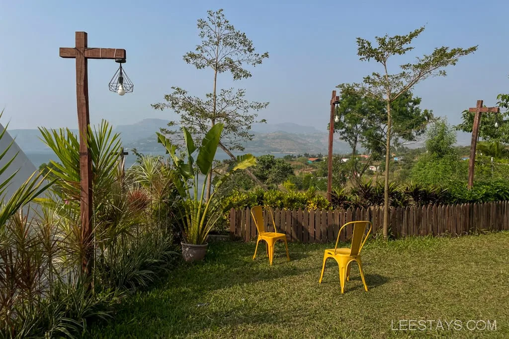 View from glass rooms near Pawna Lake, featuring yellow chairs on a lawn surrounded by plants, with wooden posts and distant hills in the background.