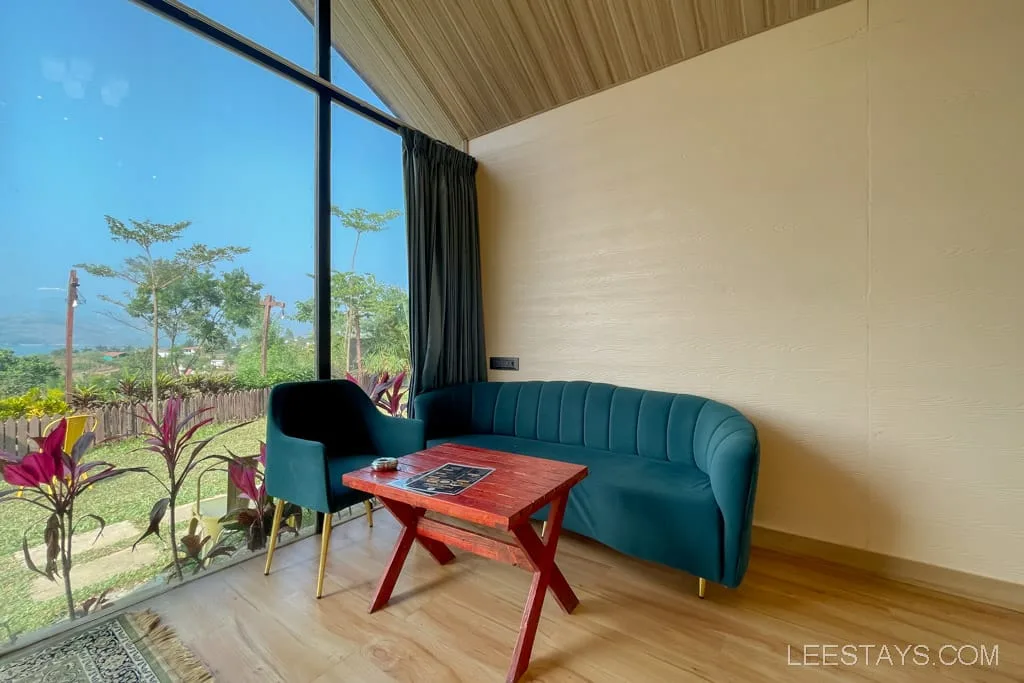 Interior of a glass room overlooking Pawna Lake, featuring a blue sofa, a black chair, and a red table surrounded by greenery outside.