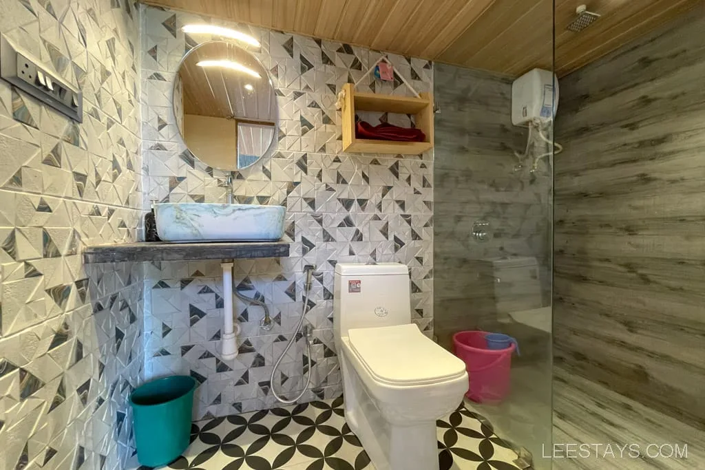 A modern bathroom featuring a geometric-patterned wall, a round mirror above a marble sink, a toilet, and a glass-enclosed shower area with a water heater. The floor has black and white geometric tiles, and there are green and pink buckets near the shower.