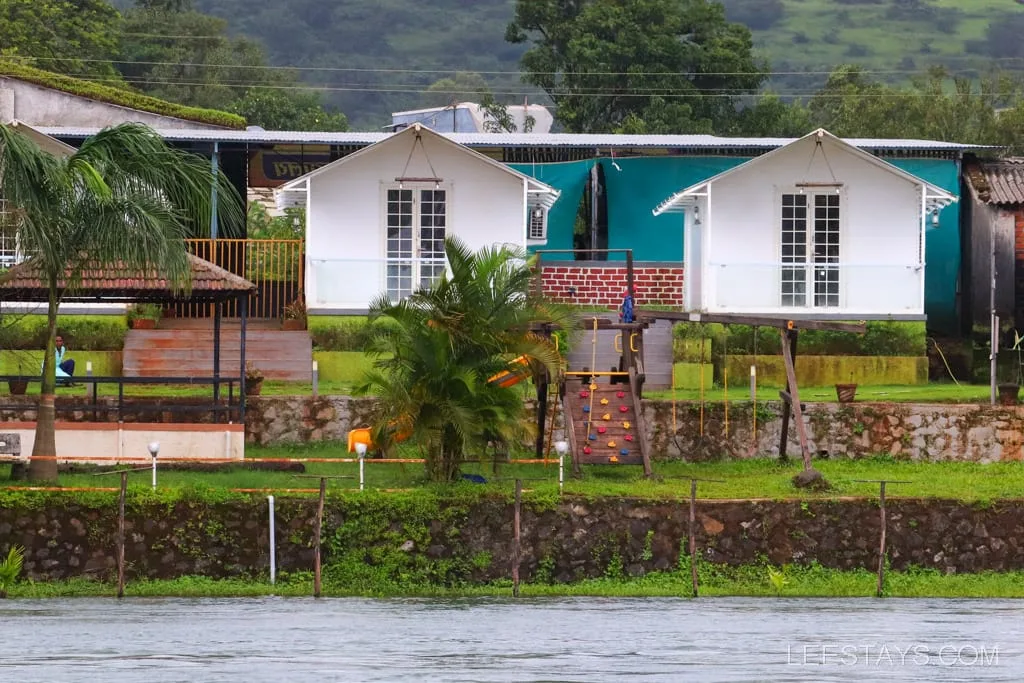 Riverside cottages by Pawna Lake with white exterior and green surroundings, featuring a palm tree and a small garden.