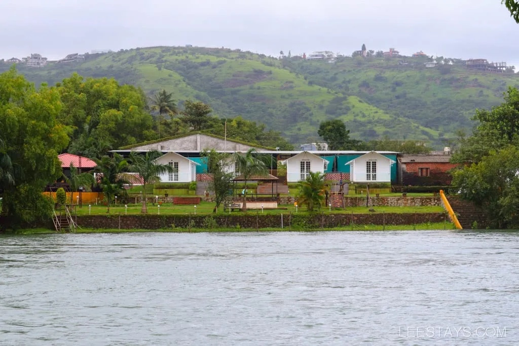 Riverside cottages by Pawna Lake, surrounded by lush greenery and rolling hills, with a body of water in the foreground.