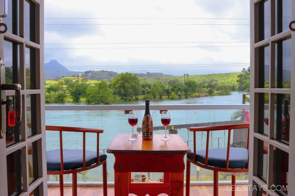 View from a riverside cottage at Pawna Lake, featuring a wooden table with two glasses of wine and a bottle, flanked by chairs, overlooking a peaceful river and green landscape.