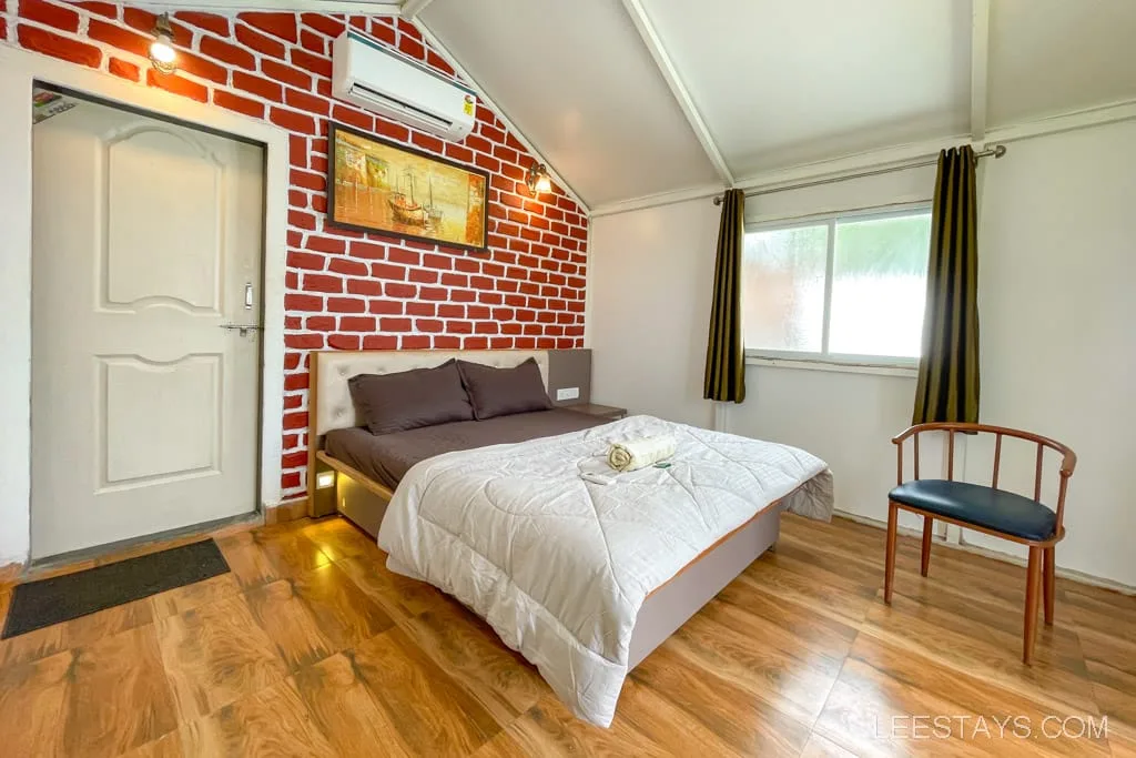 Interior of a riverside cottage near Pawna Lake featuring a cozy bedroom with a bed, wooden flooring, and a brick accent wall.