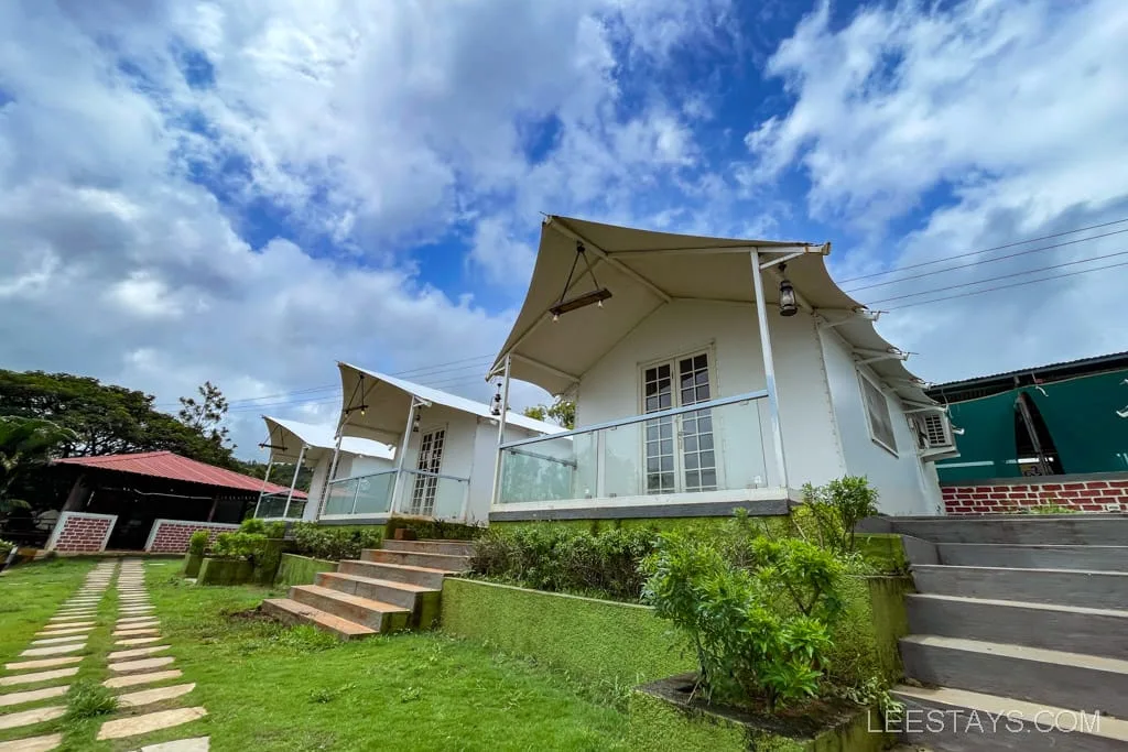 Riverside cottages near Pawna Lake with sloped canvas roofs, surrounded by greenery and a stone path under a partly cloudy sky.