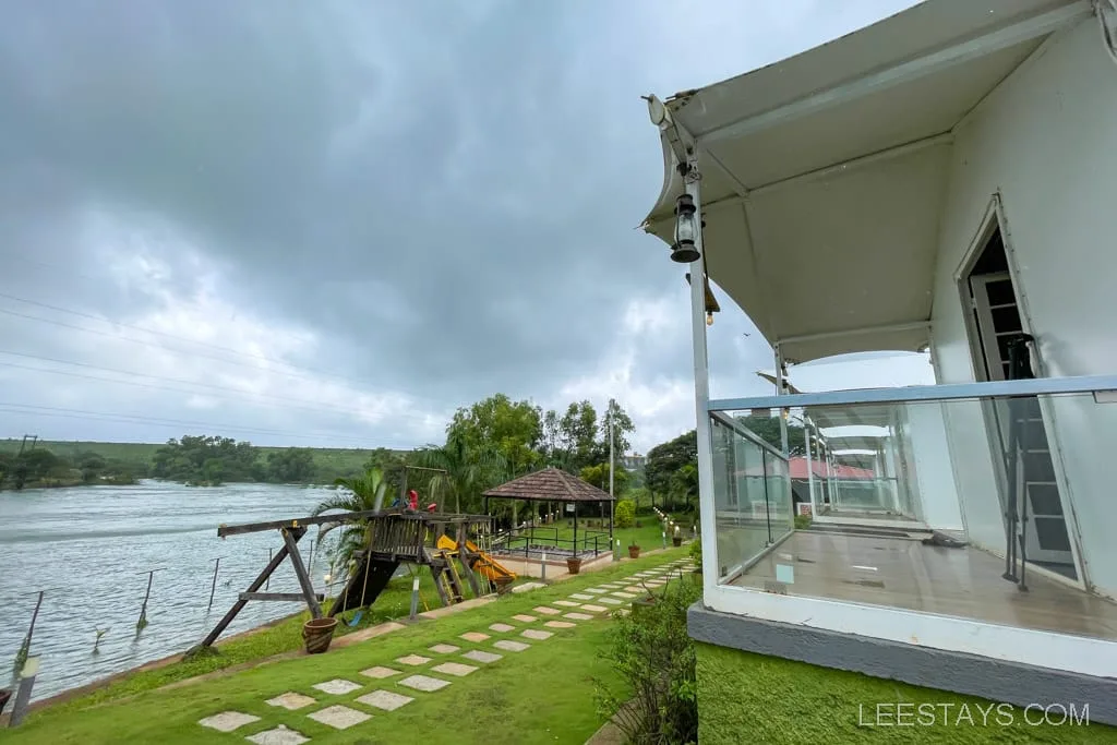 Riverside cottages at Pawna Lake with a view of the lake, a grassy pathway, and a small play area under cloudy skies.