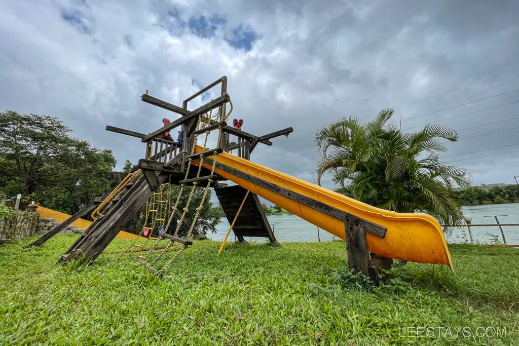A children's play area featuring a wooden structure with a yellow slide, located on the grassy grounds of riverside cottages near Pawna Lake, with a view of the lake in the background.