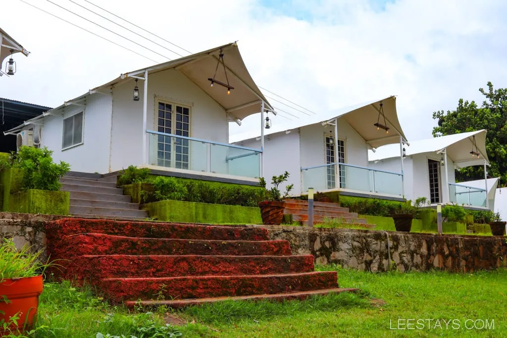A row of modern riverside cottages near Pawna Lake, featuring white exteriors, glass railings, and surrounded by lush greenery and stone steps.