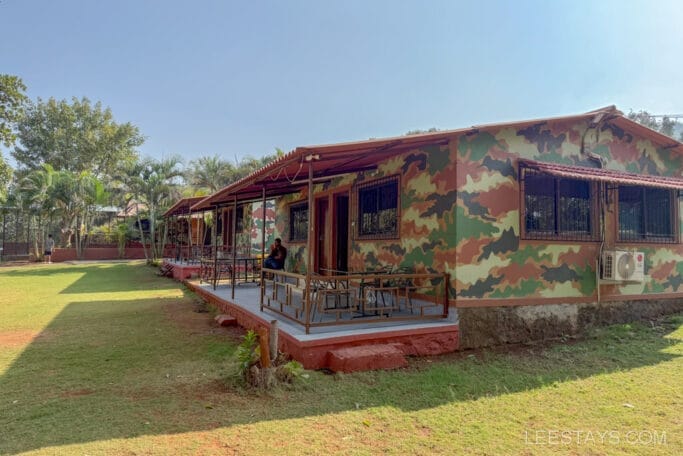 Camo-painted cabin with a porch, set in a grassy area surrounded by palm trees, ideal for a Pawna Lake stay, offering a peaceful retreat in nature.