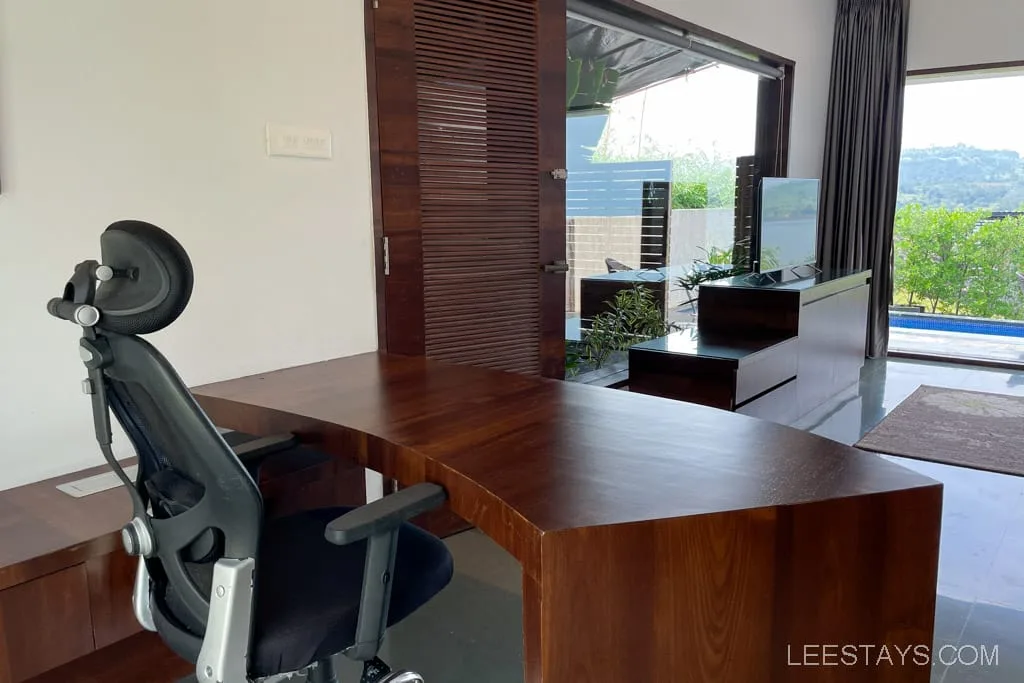 Modern resort office space near Pawna Lake featuring a curved wood desk and ergonomic chair, with views of greenery and distant hills.