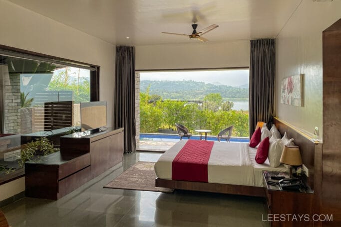 A resort room with a double bed and red accents, overlooking Pawna Lake with a view of greenery and hills through large windows.