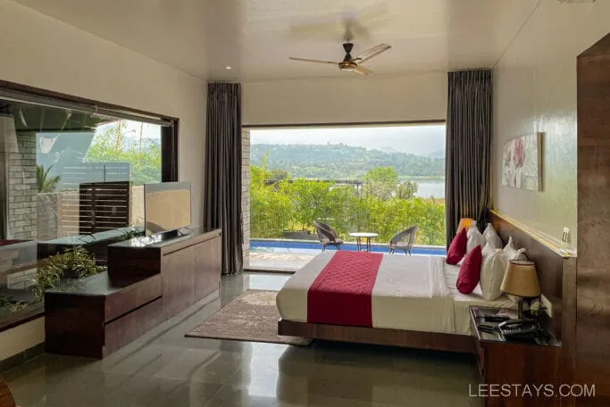 A resort room with a double bed and red accents, overlooking Pawna Lake with a view of greenery and hills through large windows.